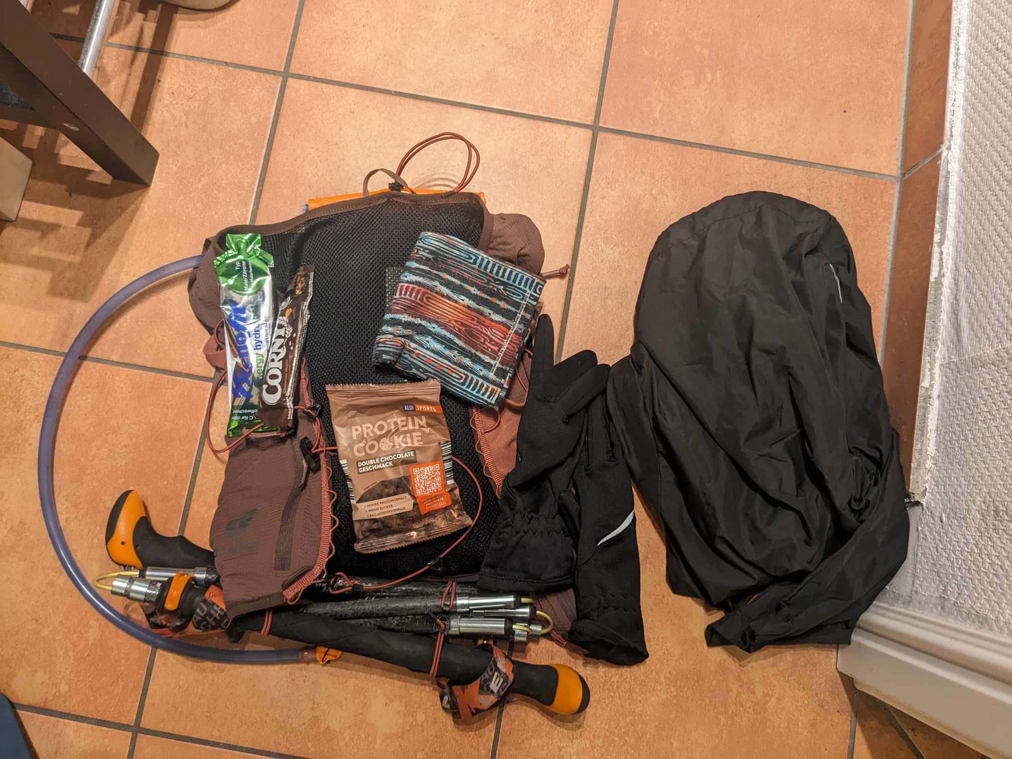 Man sieht eine Vielzahl an Ausrüstung die in den Rucksack reinpassen: Riegel, Gelbeutel, Proteinkeks, Handschuhe, Regenjacke, Trinkblase, Wanderstöcke, Buff